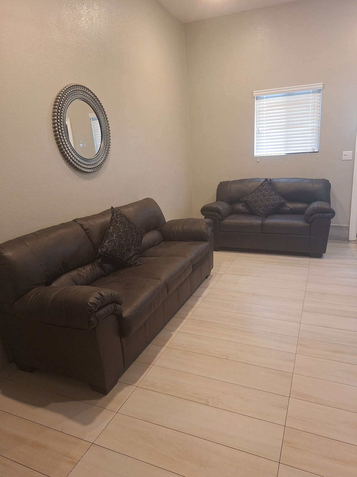 A spacious living area features two dark leather sofas, each adorned with decorative pillows. A circular mirror complements the neutral wall, while sunlight filters through a window partially covered by blinds, creating a welcoming, well-lit environment.