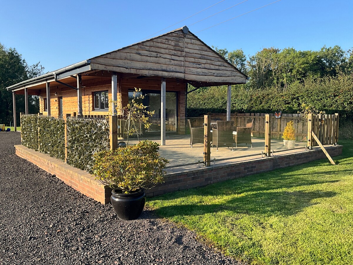 A spacious cabin is surrounded by a well-maintained garden. The wooden structure features a covered porch with multiple seating areas and a fence for privacy. Lush greenery and landscaped areas complement the outdoor space, providing a peaceful rural setting.