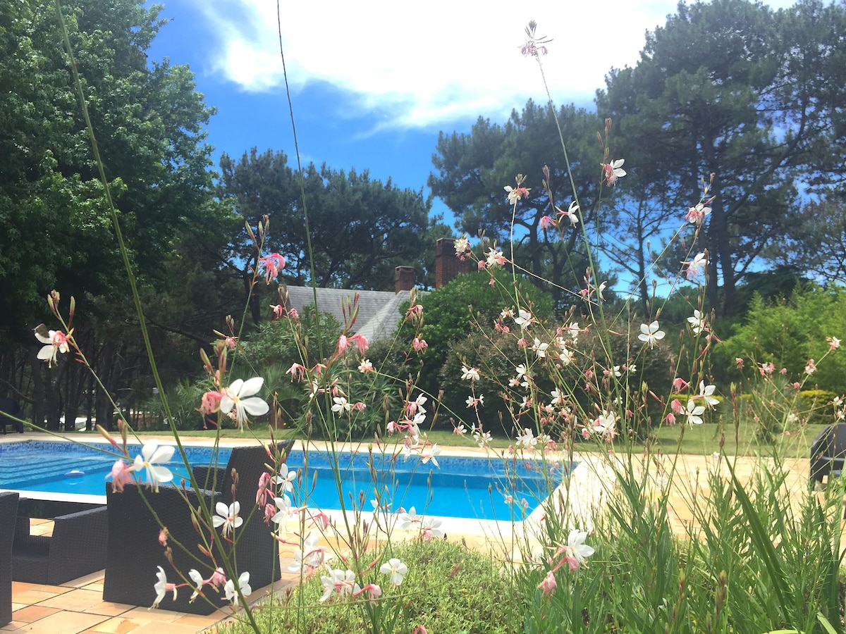 A serene outdoor area is framed by blooming pink and white flowers, leading towards a spacious swimming pool. Lush green trees in the background enhance the natural setting, while a cozy seating arrangement is positioned nearby for relaxation.