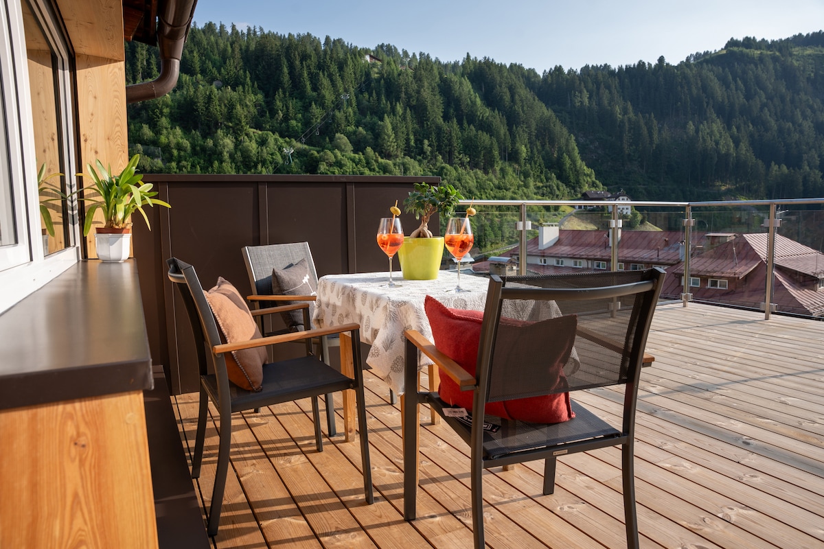 A spacious wooden terrace is featured, furnished with a table surrounded by four chairs. A large plant adds greenery, while two glasses and a vibrant yellow planter provide color. The backdrop includes lush green hills and distant buildings, creating a serene outdoor setting.