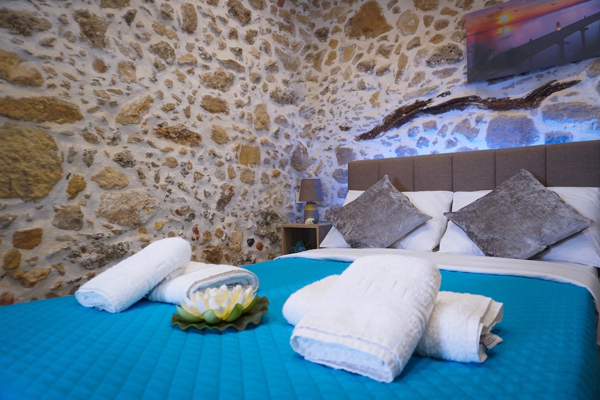 The room features a textured stone wall, providing a rustic ambiance. A neatly made bed with blue bedding is centered, adorned with folded towels and a decorative item. Soft lighting from a bedside lamp enhances the serene atmosphere.