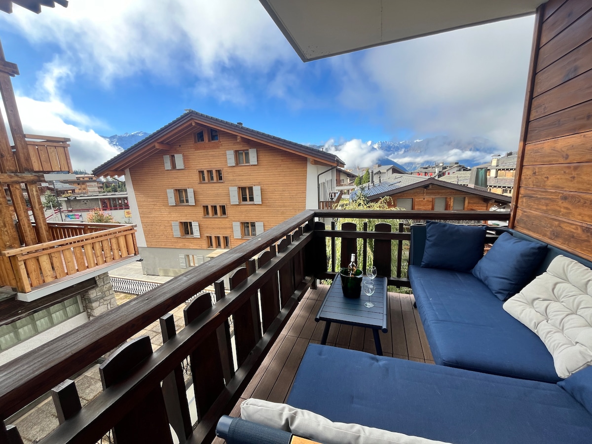 A covered terrace features a seating area with plush cushions and a low table. The surrounding wooden railing offers a view of nearby buildings and expansive mountainous landscapes under a cloudy sky.
