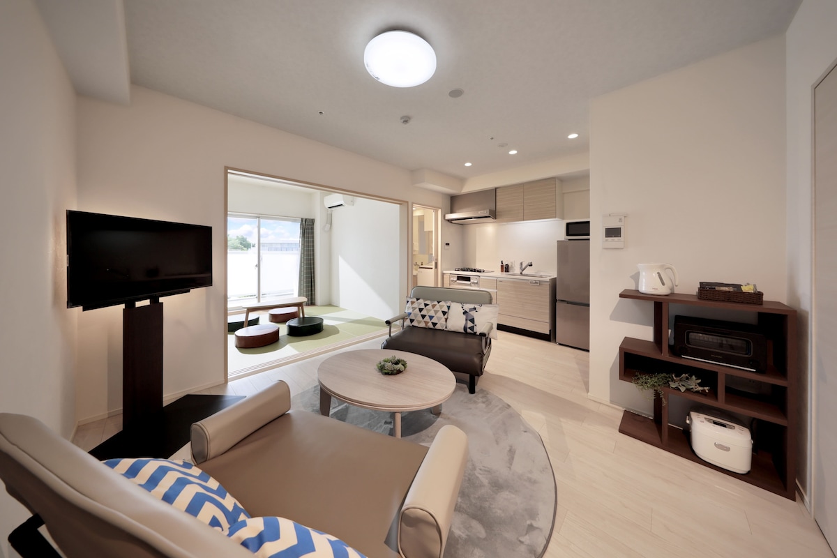 A spacious living area is showcased, featuring a comfortable seating arrangement with patterned cushions. A flat-screen TV is positioned on a stand. The open layout leads to a kitchen area with modern appliances, enhancing the inviting space bathed in natural light.