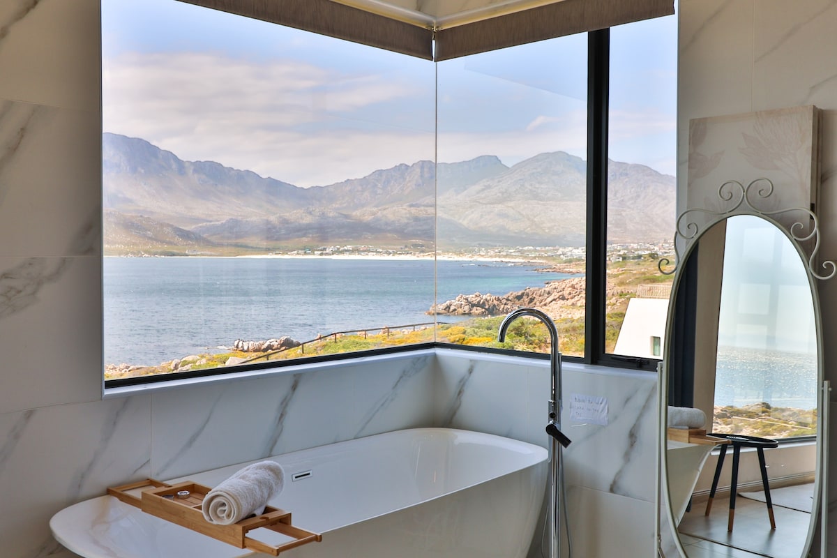 A modern bathroom features a freestanding tub positioned beside a large window. Breathtaking views of the ocean and mountains are visible, framed by soft natural light. A wooden tray rests on the edge of the tub, complementing the elegant marble design.
