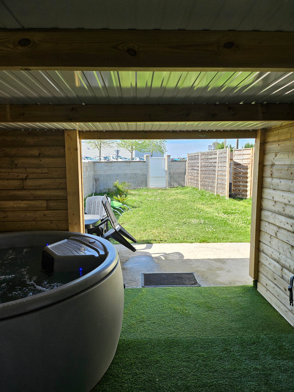 A view from inside a wooden structure shows a jacuzzi with bubbling water beside a small outdoor space. Green grass and a path lead to a gate, while a chair rests nearby. Bright light enters through the open door, creating a sense of openness.