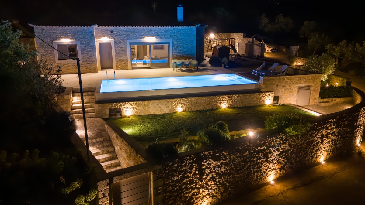 The stone house is illuminated at night, showcasing a modern pool with a clear blue surface. Soft lighting highlights the surrounding walls and steps, while lounge chairs are positioned nearby. The mountain backdrop adds a serene ambiance to the outdoor space.