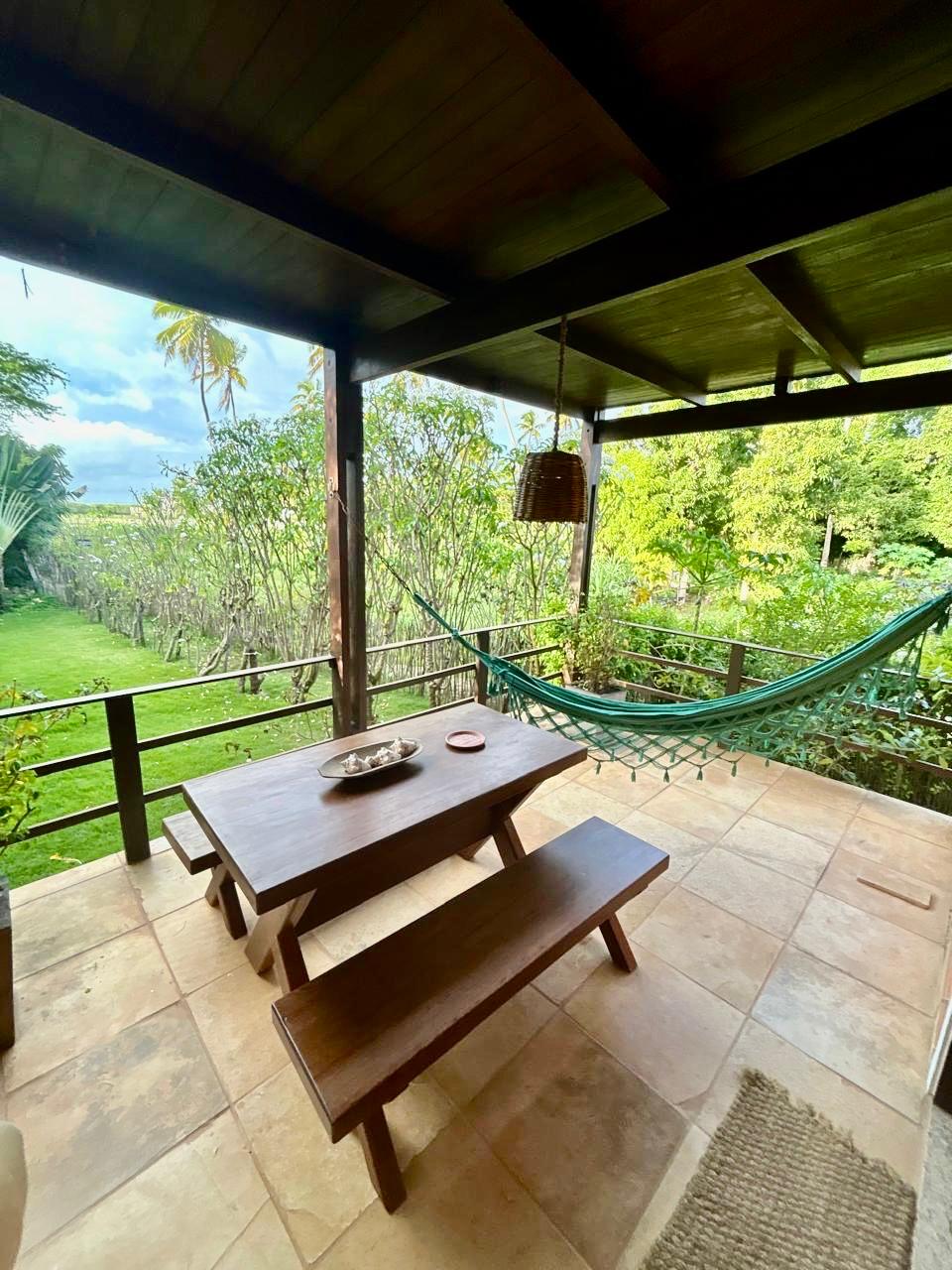 A covered outdoor area features a wooden table and benches, providing an inviting space for dining. A hammock is suspended nearby, allowing for relaxation while enjoying the surrounding greenery. Lush plants and trees are visible just beyond the railing, enhancing the tranquil atmosphere.