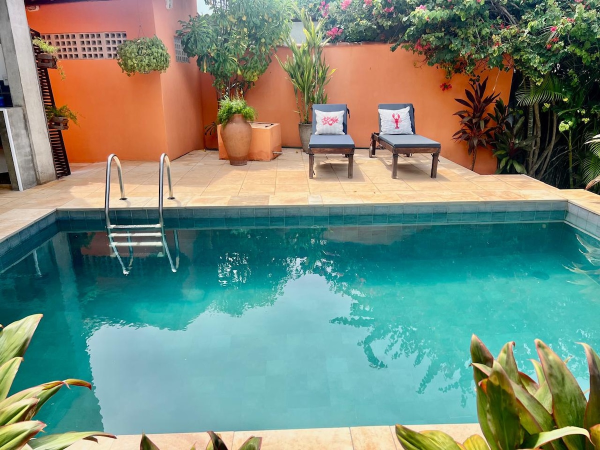 A serene swimming pool is framed by lush greenery and colorful plants. Two sun loungers with soft cushions are positioned nearby, creating a relaxing spot. An accent wall in warm tones adds to the inviting atmosphere, while steps lead into the clear water.