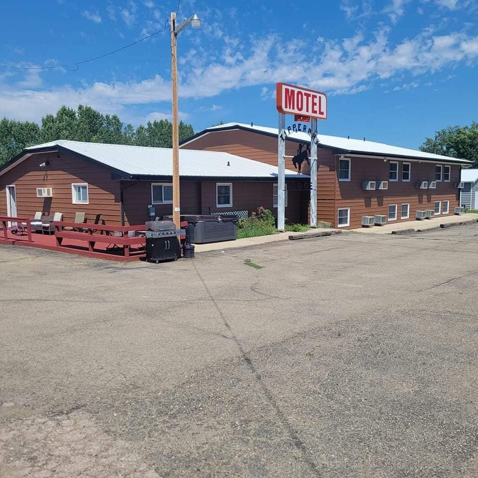 The exterior of the Tipperary Motel & Lodge features a wooden façade and a prominent sign displaying the motel's name. A wide gravel path leads to the entrance, with a seated area in front, surrounded by trees under a clear blue sky.