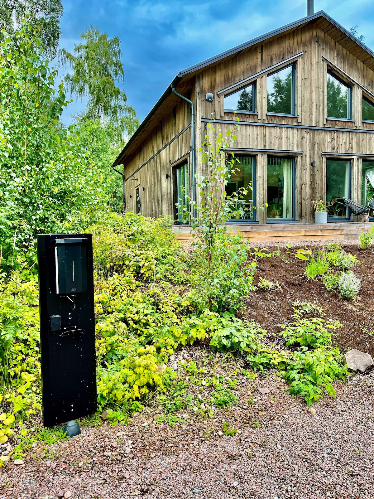 The modern villa features large glass windows that invite natural light, surrounded by lush greenery. A charging station for electric vehicles is positioned prominently in the foreground, highlighting the property's eco-friendly amenities. The warm tones of the wooden exterior complement the natural setting.
