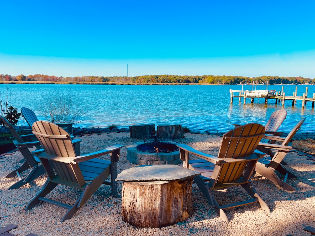 A circular arrangement of five wooden Adirondack chairs surrounds a fire pit on a sandy shoreline. The calm water reflects a clear blue sky, with a view of lush trees lining the opposite bank. A wooden pier extends into the water in the background.