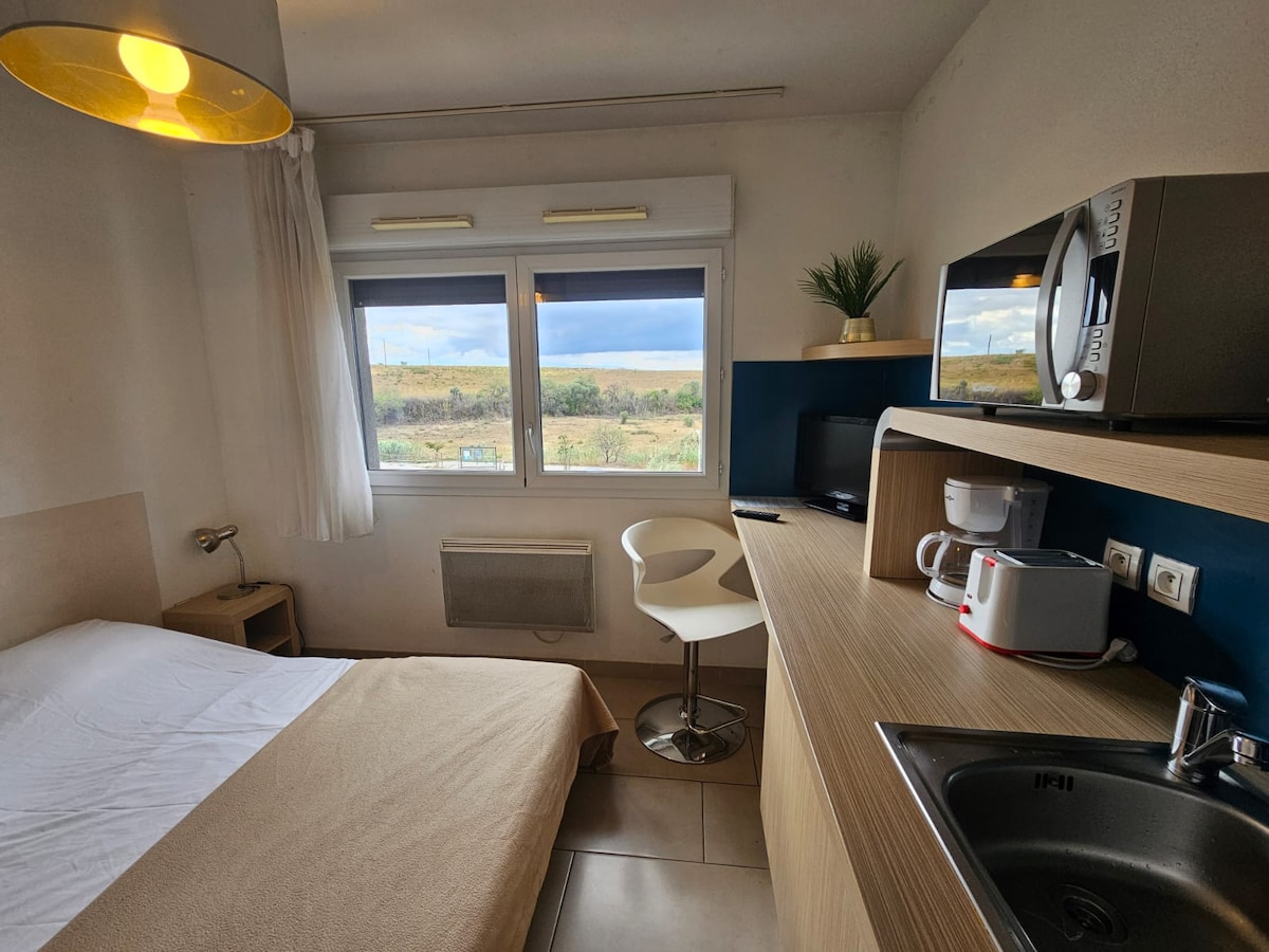 A cozy studio features a spacious bed with a neutral-toned blanket. A modern kitchenette with a microwave and coffee maker is set against a blue accent wall. A small dining area includes a white chair and table. Large windows offer a view of the surrounding landscape.