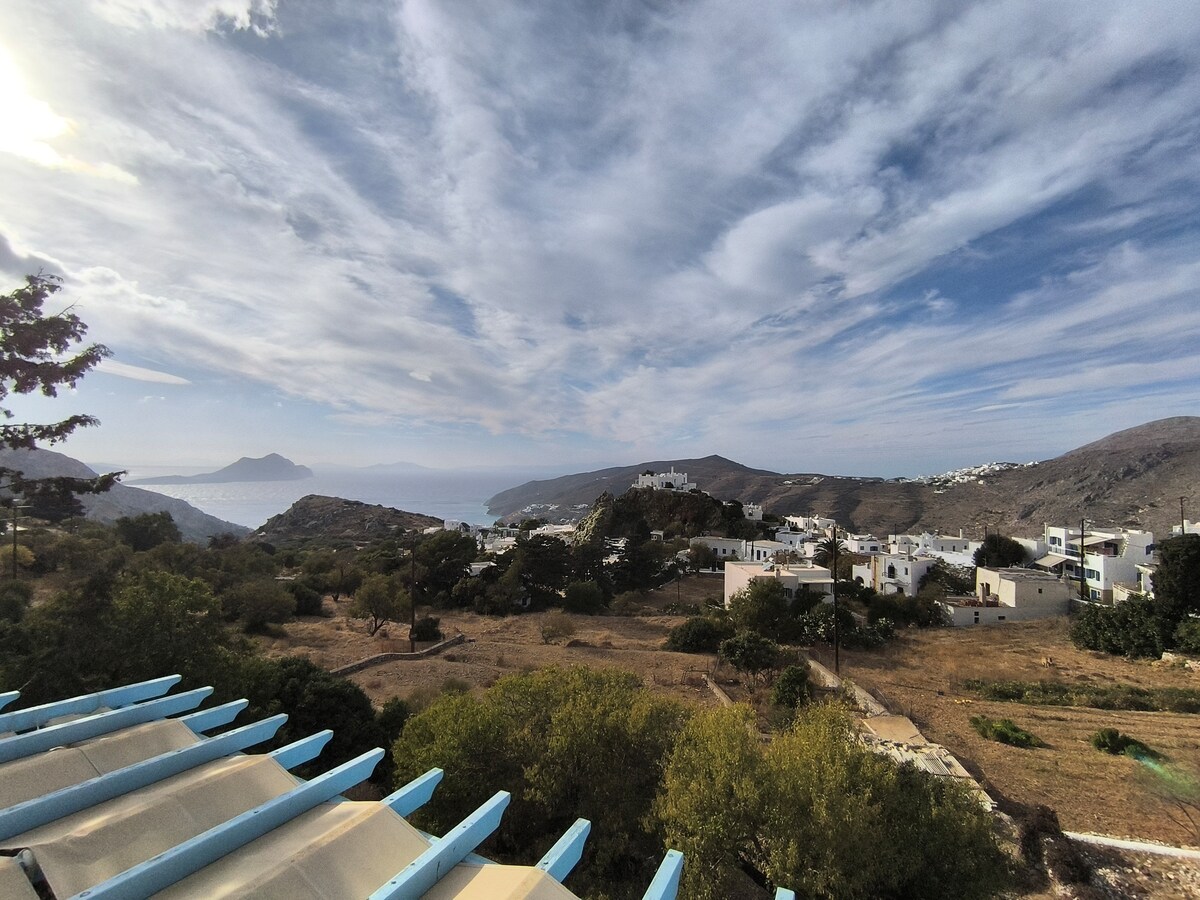 A panoramic view captures the serene landscape of Lagkada, featuring rolling hills and whitewashed buildings. The coastline and distant mountains create a harmonious blend of nature and habitation, while fluffy clouds drift in the expansive sky above.
