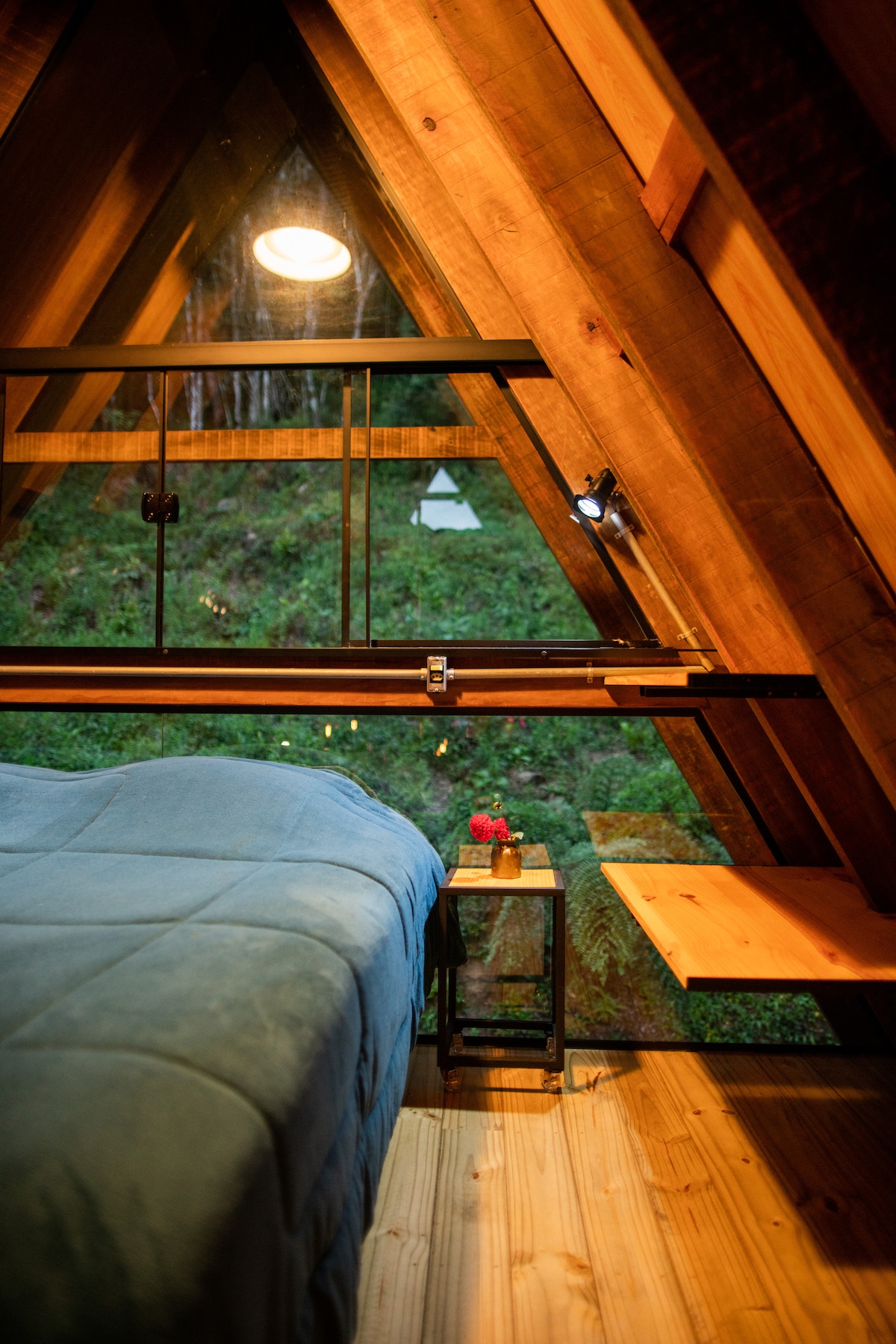 The triangular interior features wooden beams and a large window that frames views of lush greenery. A spacious bed is adorned with a soft blue cover, complemented by a bedside table holding a small flower arrangement. A wooden shelf extends along the wall, adding functionality.