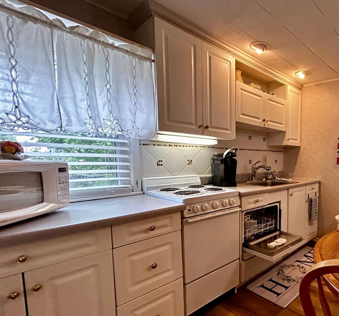 A well-equipped kitchen features white cabinetry and appliances, including a microwave, stove, and dishwasher. An elegant countertop provides ample workspace. Natural light streams in through a window adorned with a sheer curtain, enhancing the bright ambiance of the room.