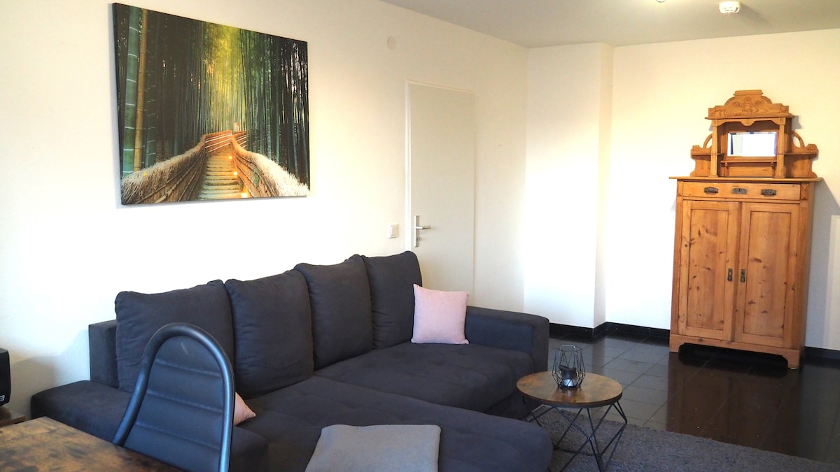 A spacious living area is highlighted by a comfortable dark sofa adorned with light cushions. A wooden cabinet stands against the wall, complemented by a vibrant forest-themed artwork. Natural light enters through a nearby window, enhancing the room's inviting atmosphere.