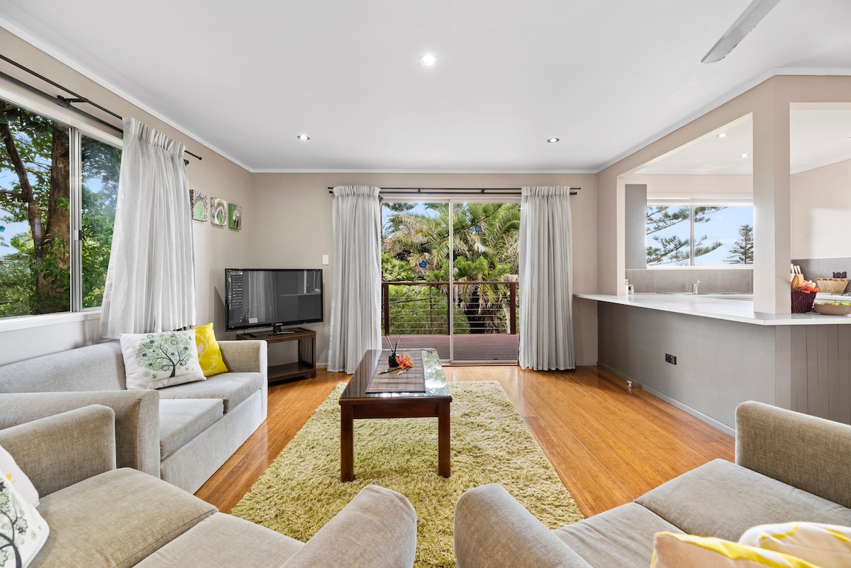 A spacious living area features light wood flooring and large windows, allowing natural light to illuminate the space. Two comfortable sofas and a coffee table are centered around the room, with a television present. A seamless transition to an outdoor balcony can be observed through the glass doors.