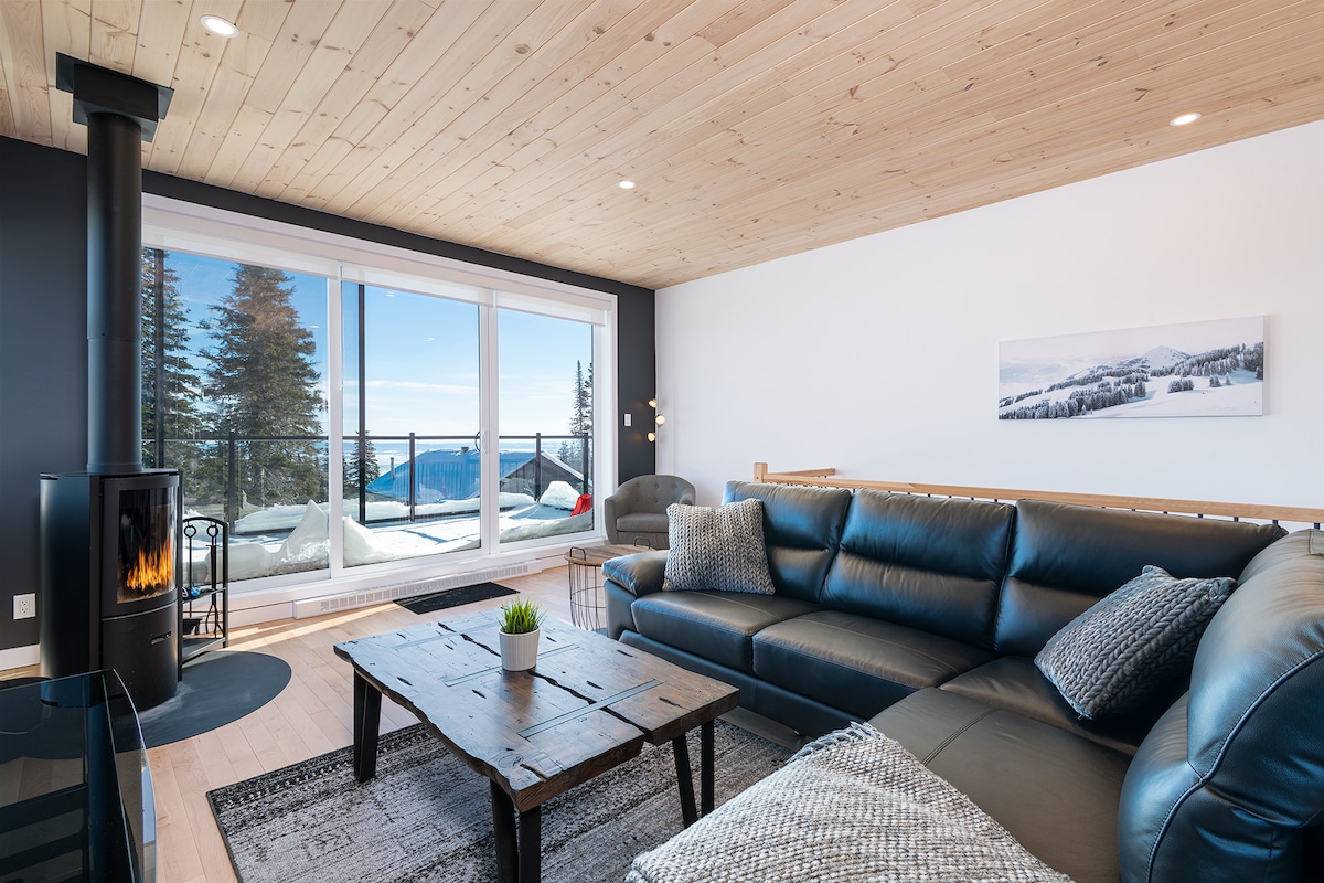 A modern living area features a dark leather sectional sofa paired with a rustic wooden coffee table. A sleek stove stands against a wall of light wood paneling. Large glass doors reveal a scenic view of the outdoors, with trees and mountains visible beyond.