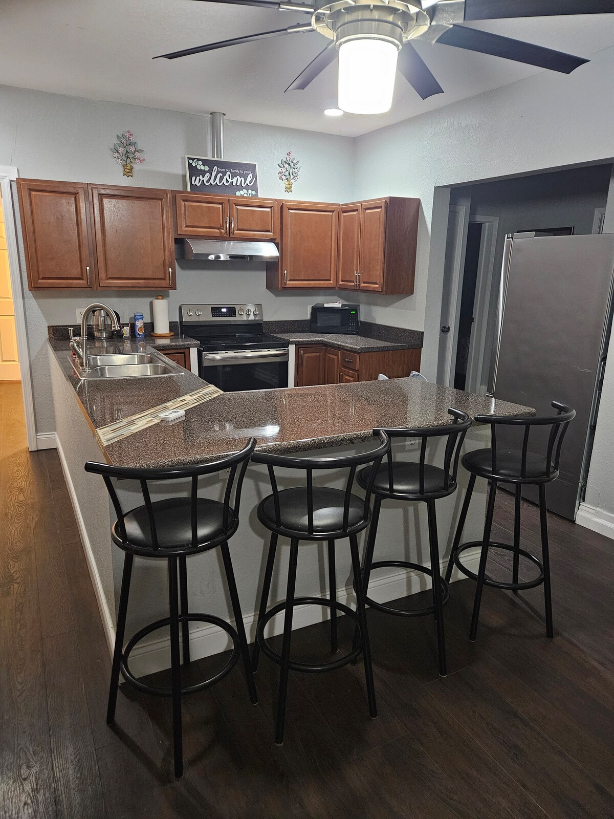 A modern kitchen features wooden cabinets and a granite countertop, complemented by a four-stool bar area. Stainless steel appliances, including an oven and refrigerator, are integrated into the design. The space is illuminated by overhead lighting, enhancing a bright and functional ambiance.