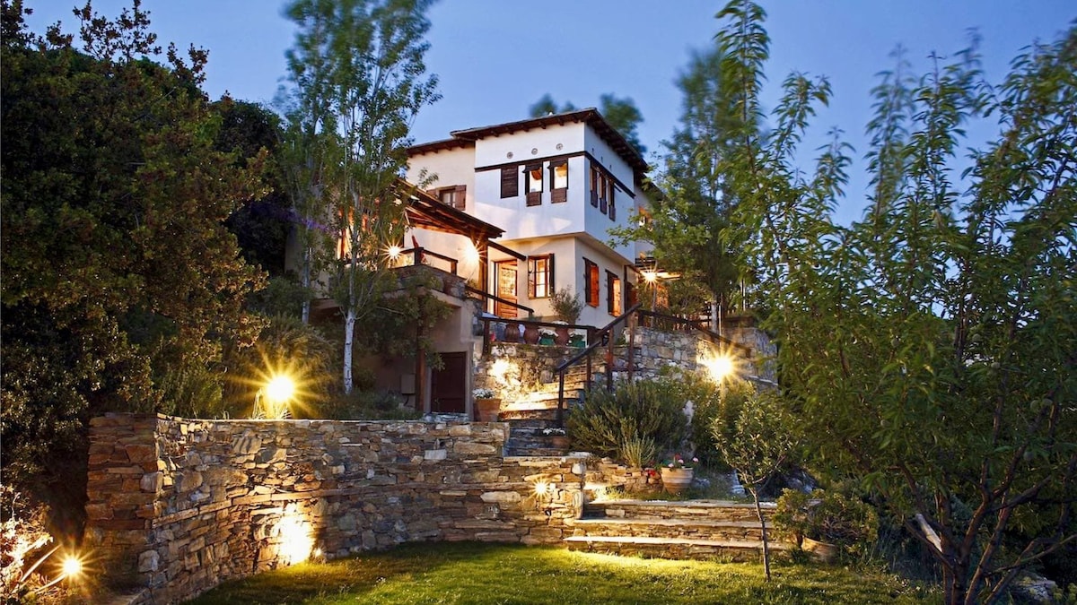 A two-story house is set amidst lush greenery, illuminated by soft outdoor lighting. Stone steps lead up to the entrance, surrounded by various trees. The design features wooden balconies and large windows, offering a blend of indoor comfort and outdoor charm.