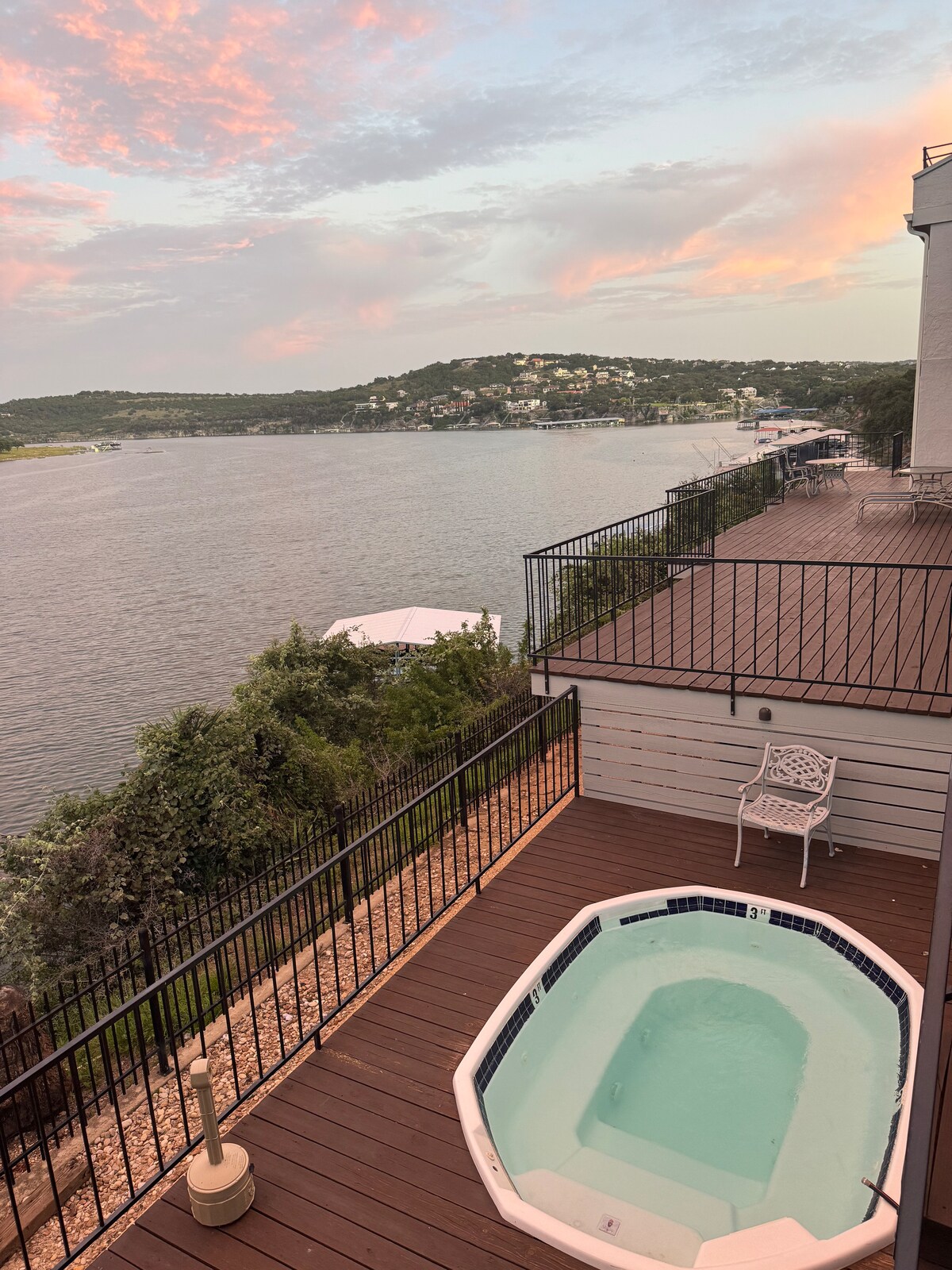 A private hot tub is positioned on a wooden deck, overlooking the serene waters of Lake Travis. Lush greenery surrounds the area, while distant hills form a picturesque backdrop against the softly colored sky at sunset.