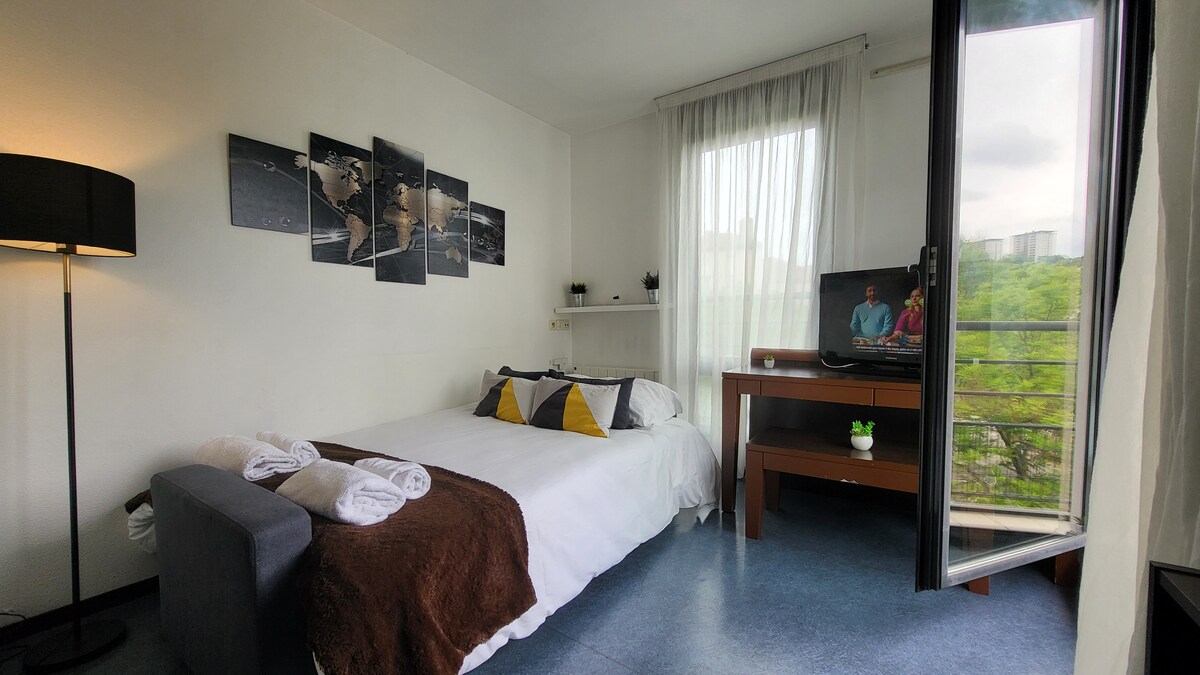 A cozy sleeping area features a spacious bed with a neutral-coloured blanket and decorative pillows. A TV is positioned on a wooden console, while a lamp provides additional lighting. The open balcony allows natural light to brighten the room and offers views of greenery outside.