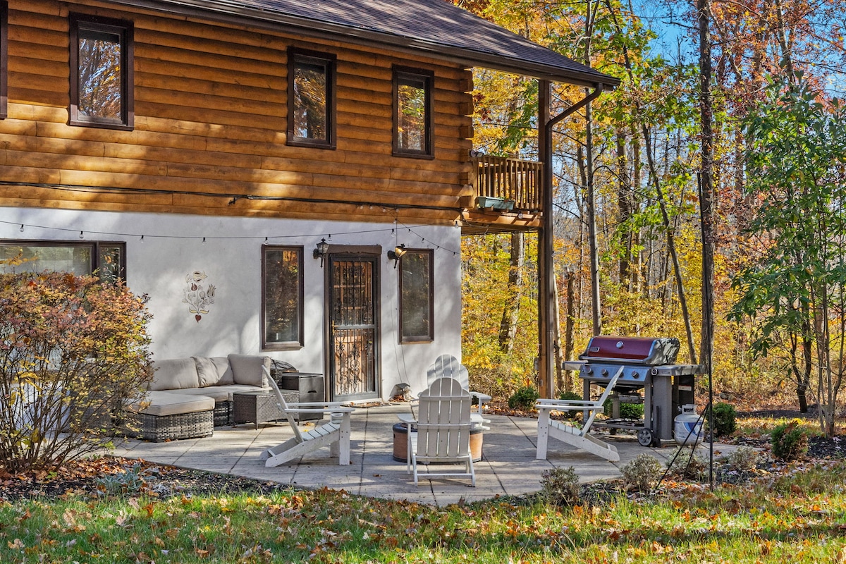 The outdoor patio is surrounded by trees, featuring a cozy seating area with a sectional couch and several lounge chairs. A fire pit is situated in the center, and a gas grill is positioned nearby, inviting outdoor gatherings.