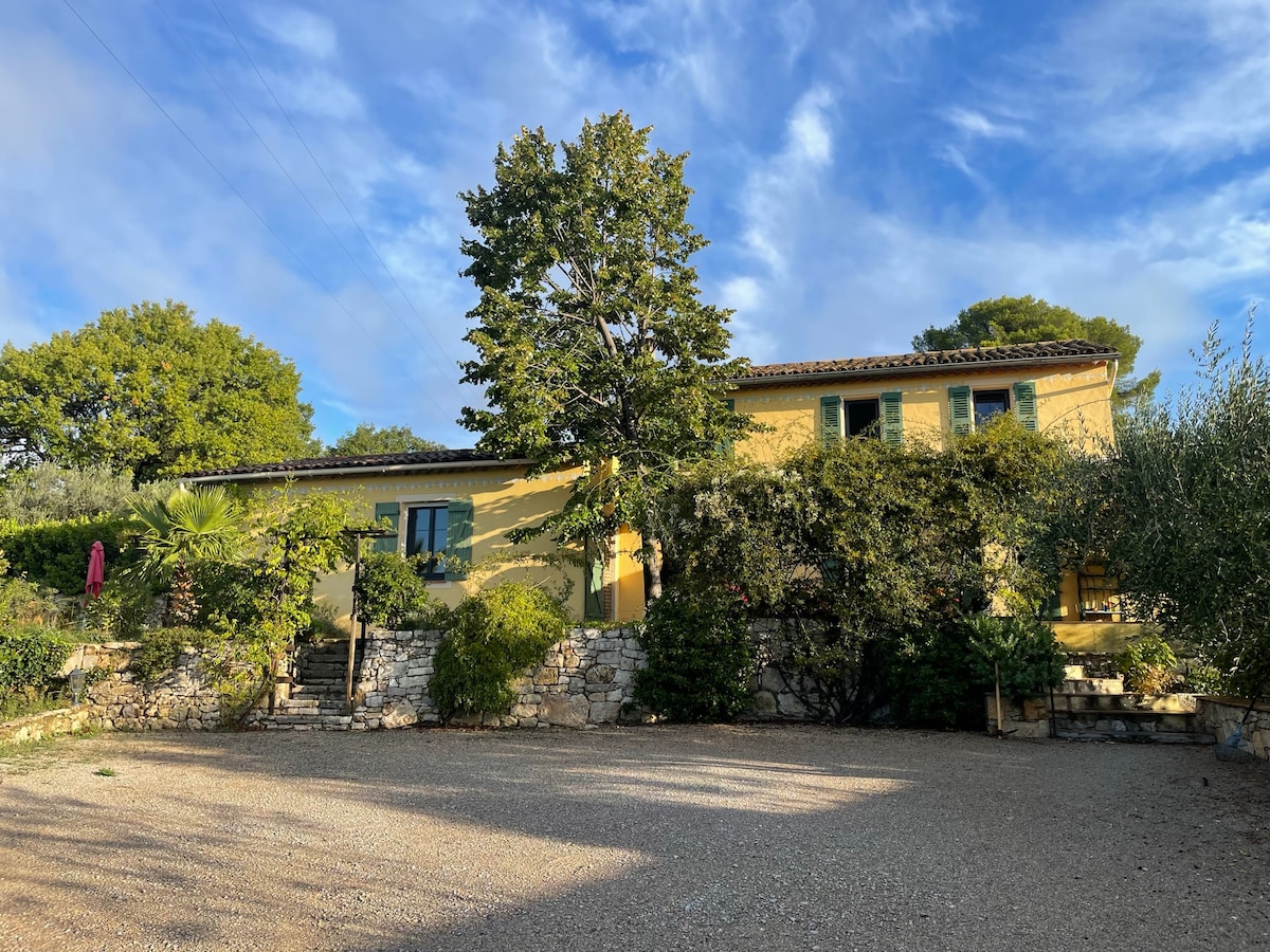 A charming yellow villa is framed by lush greenery and a variety of mature trees. The stone wall adds character to the exterior. A gravel parking area leads up to the entrance, showcasing the spacious outdoor setting, with a sunny sky in the background.