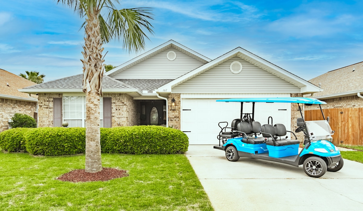 The exterior of the home features a well-manicured lawn with a palm tree and vibrant green shrubs. A blue and white golf cart is parked in the driveway, ready for use. The house is a one-story structure with a brick facade and a welcoming entrance.