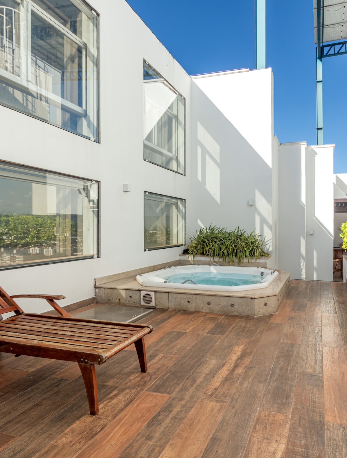A private outdoor area features a jacuzzi surrounded by tiled flooring. Sunlight filters through large windows, casting natural light. A wooden lounge chair is positioned nearby for relaxation.