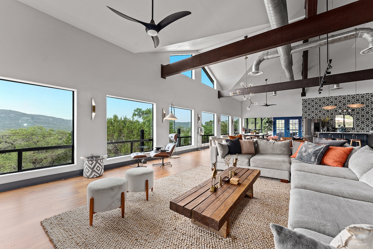 A bright, open-concept living area features large windows offering views of the surrounding hills. A plush sectional sofa is arranged around a rustic wooden coffee table, complemented by two small upholstered stools. High ceilings and unique lighting fixtures enhance the spacious atmosphere.
