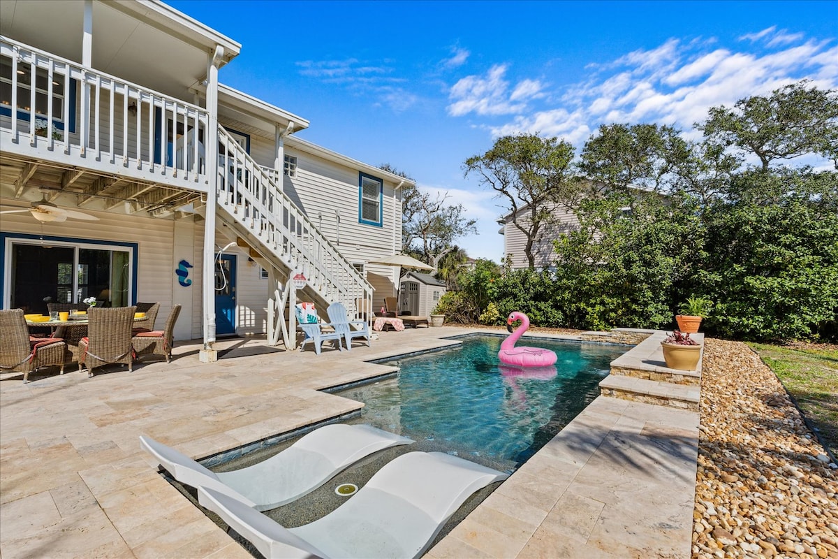 The outdoor space features a private saltwater pool with a fountain and lounge chairs. A pink flamingo float rests in the water. The area is surrounded by landscaping and has multiple seating arrangements, including a dining table set on a stone patio beneath the deck.