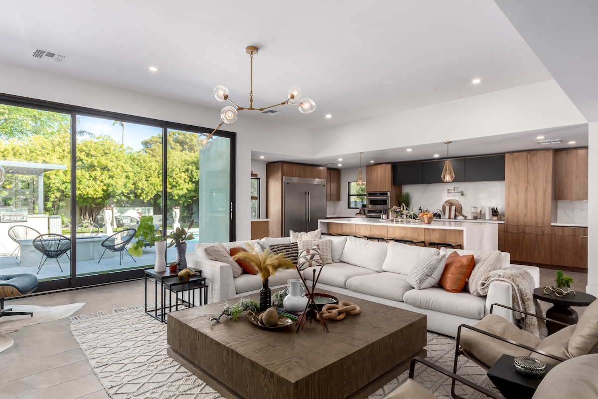An open living area features a spacious sectional sofa with decorative pillows, surrounded by natural light from large glass doors. A wooden coffee table sits at the center, adorned with plants and decorative items, seamlessly connecting to the kitchen and outdoor space.