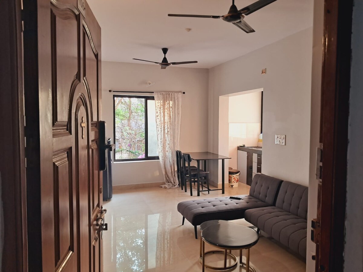 An entryway reveals a living area furnished with a dark sectional couch and a circular coffee table. A dining table with chairs is positioned nearby. Large windows allow natural light to fill the space, complemented by light-colored curtains and a neutral wall palette.