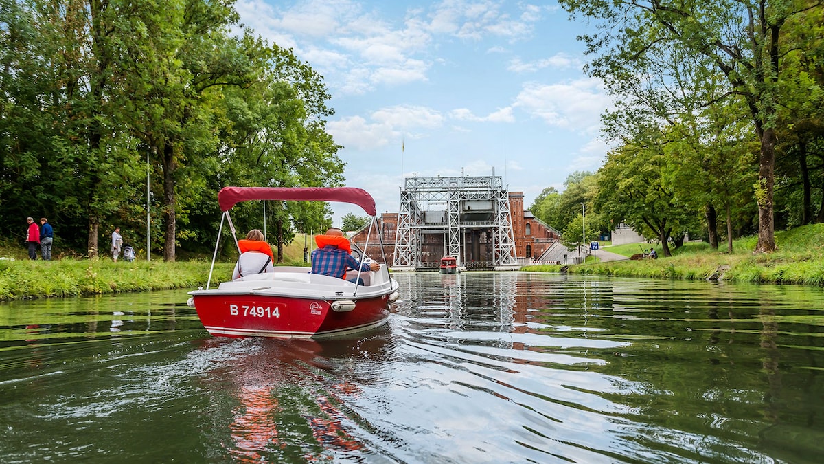 A small motorboat glides along a calm waterway, framed by lush greenery. A gentle slope leads to a historic lock structure in the background, where trees provide shade and enhance the serene environment. A few people can be seen casually strolling near the bank.