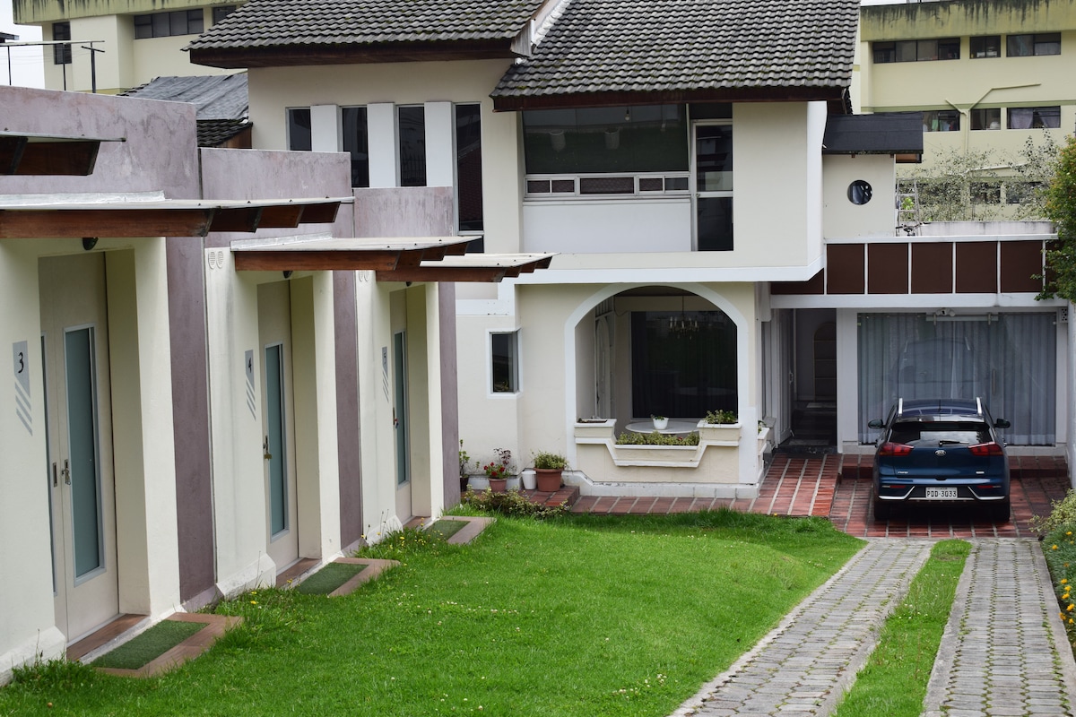 The exterior view of the property shows a well-maintained lawn leading to two separate structures. One structure features individual entrance doors, while another larger building displays a welcoming porch area. A parked vehicle is positioned in the driveway, illustrating easy access.