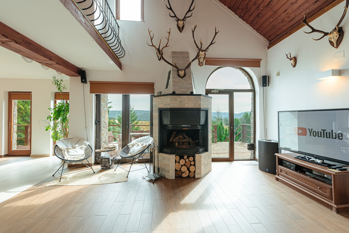 A spacious living area features a central stone fireplace, framed by exposed wooden beams and adorned with mounted antlers. Large windows provide scenic views, while comfortable seating arrangements invite relaxation. A television is positioned nearby, enhancing the entertainment options in this cozy space.