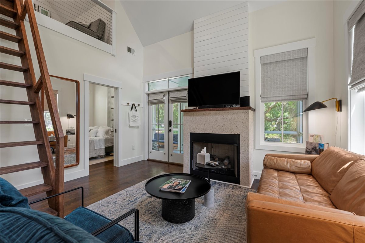 A living area features a modern leather sofa and a black coffee table, with a flat-screen TV mounted above a stone fireplace. Large windows allow natural light to fill the space, while a staircase leads to a loft area. Neutral tones create a calming ambiance.