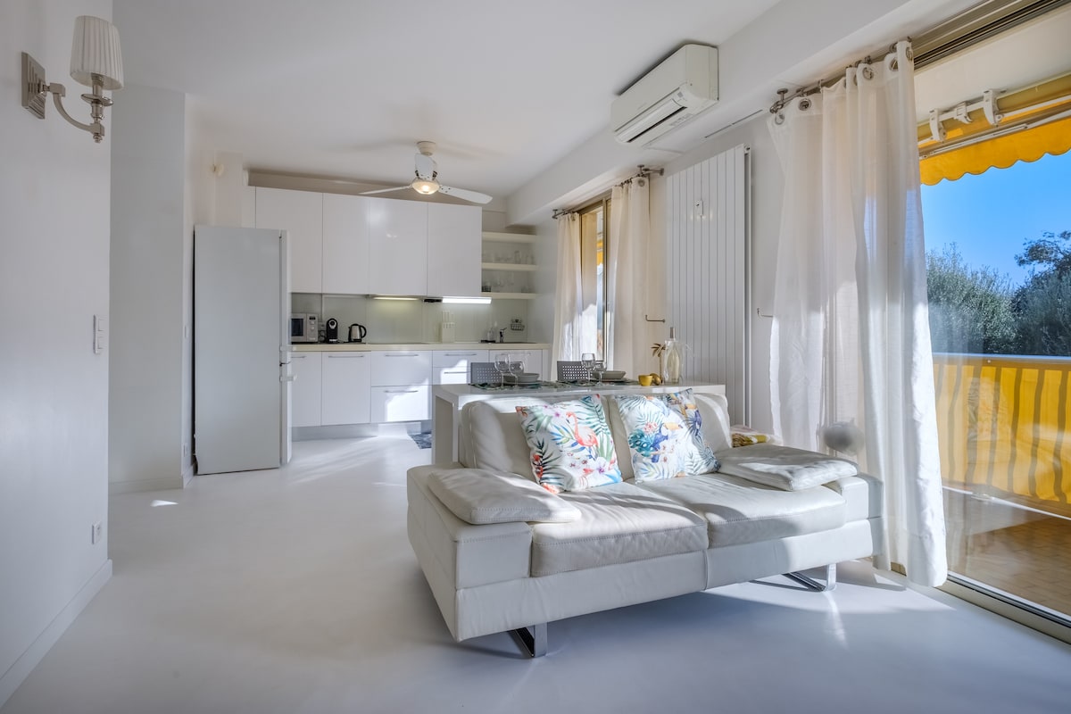 A bright living area features a comfortable white leather sofa adorned with colorful cushions. The modern kitchen is visible, equipped with essential appliances. Large sliding doors lead to a balcony with visibility of greenery, allowing natural light to fill the space.