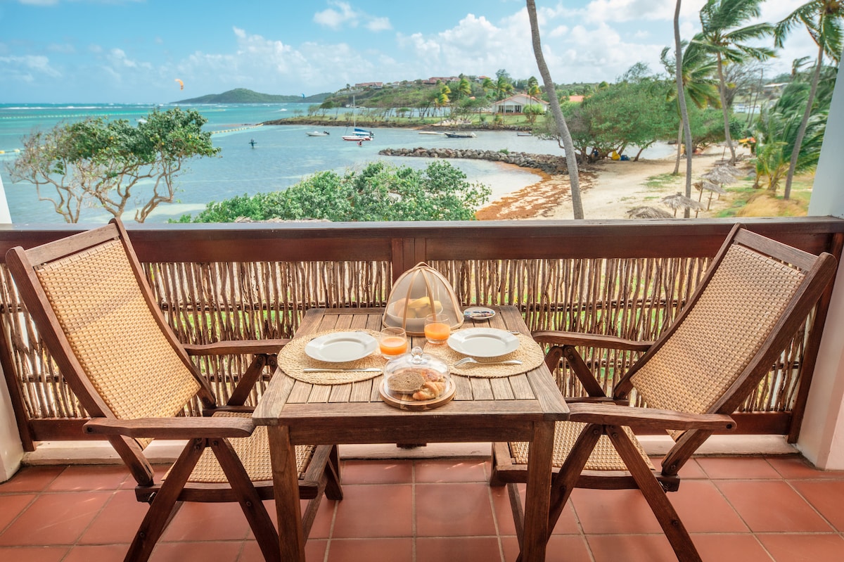 A wooden balcony table is set for two, featuring two plates and glasses. Two wicker chairs are positioned around the table. In the background, a scenic view of turquoise waters and a sandy beach is visible, framed by palm trees and distant greenery.