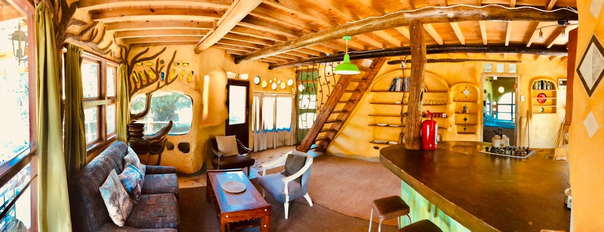An open-plan living space features textured cob walls with artistic details. Natural wood beams line the ceiling, and large windows allow ample light. A bar counter and lounge area are present, alongside a staircase leading to the upper floor. Various shelves line the walls, enhancing the rustic design.