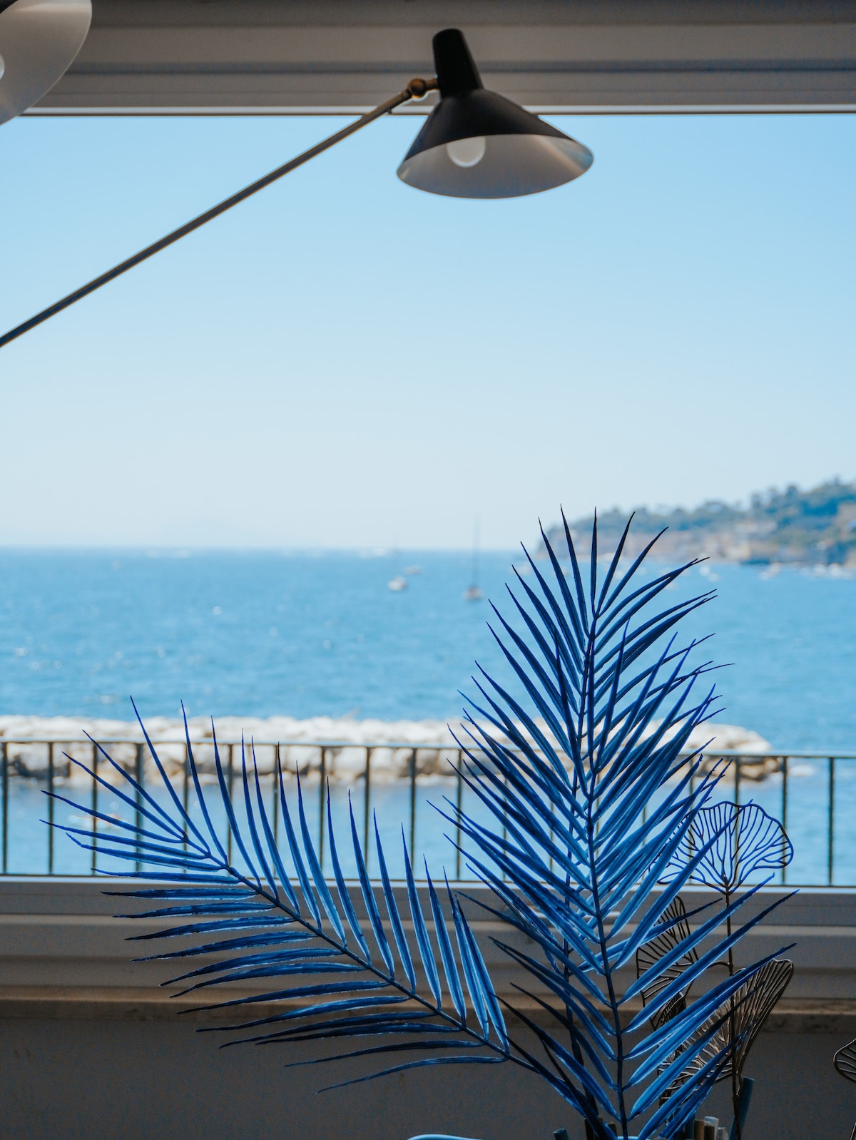 A view of the blue sea is captured through a large window, framed by a stylish lamp and decorative foliage. The sunlight reflects off the water, enhancing the tranquil atmosphere of the coastal scenery.