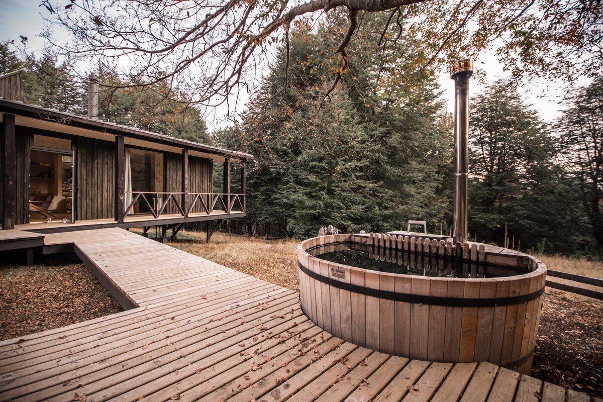 A wooden deck surrounds a round hot tub, inviting relaxation. The rustic exterior of the house features large glass doors, revealing glimpses of the interior space. Nearby, native trees provide a natural backdrop, enhancing the tranquil setting.