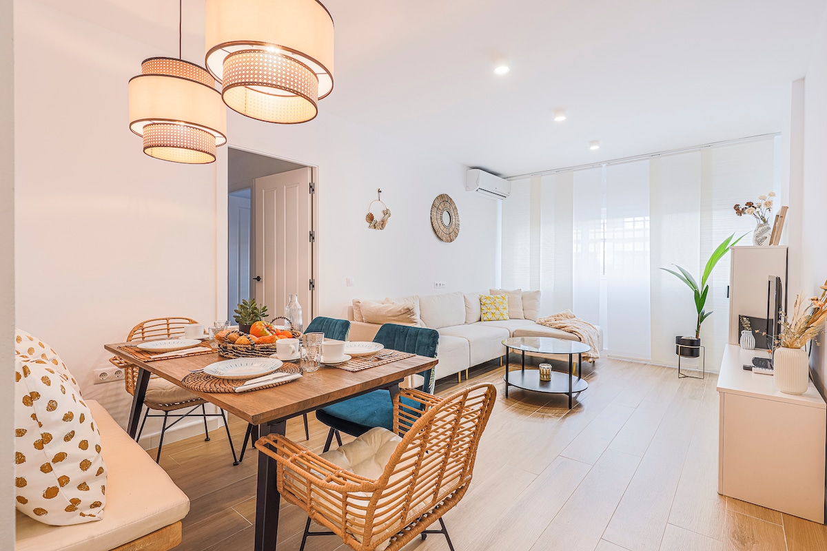 A bright living area features a dining table set for four next to a comfortable seating arrangement. Pendant lights hang above, and a coffee table complements the space. Large windows provide ample natural light, with indoor plants adding a touch of greenery.