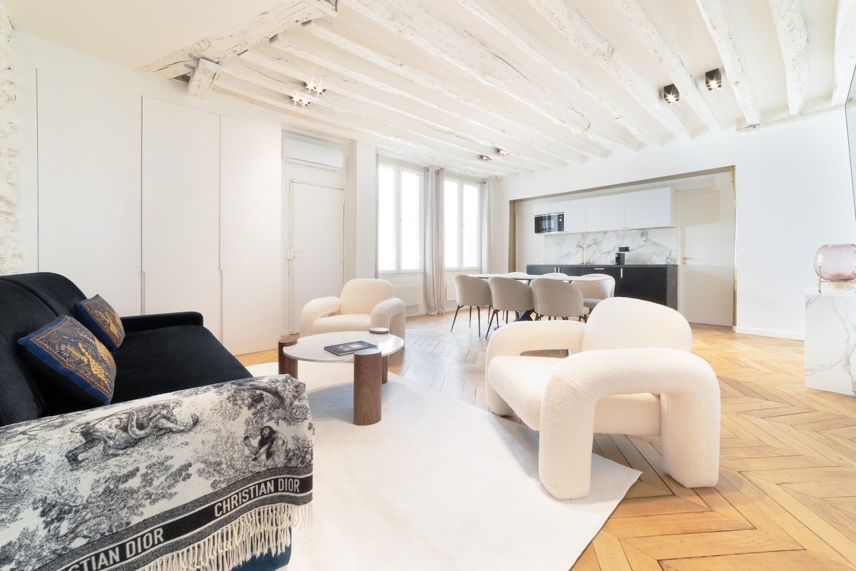 The open living area features a dark sofa adorned with a decorative throw, a round coffee table, and a light area rug. Large windows allow natural light to fill the space, which includes a dining setup for six and a modern kitchen with marble accents.