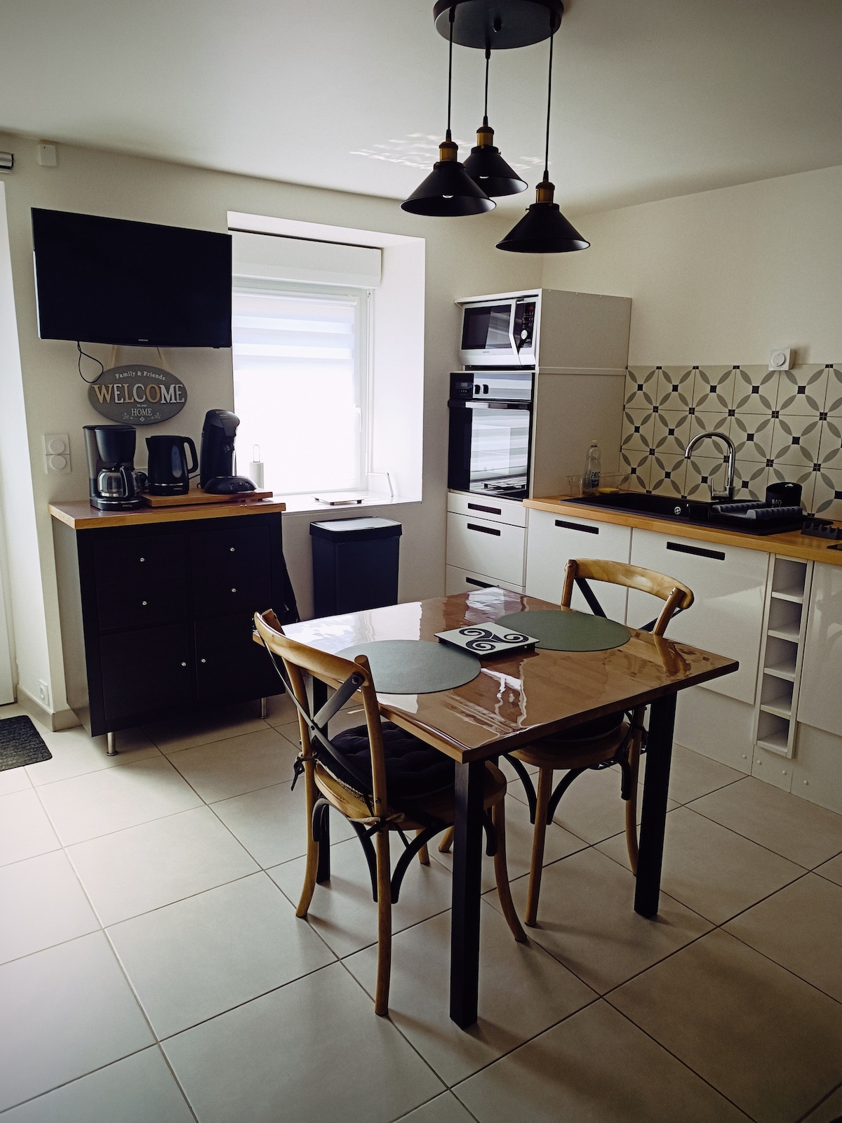 A modern kitchen area is presented, featuring sleek cabinetry and a dining table with four wooden chairs. Appliances including a coffee maker and microwave are visible. Natural light enters through a window, highlighting the contemporary decor and tile backsplash.