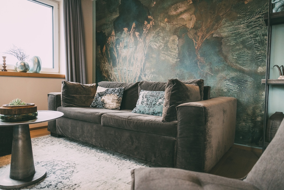 A comfortable living area features a plush gray sofa adorned with decorative cushions. A small round table sits nearby, with a decorative centerpiece placed on top. A textured wall art piece, depicting nature, adds a serene backdrop to the space.