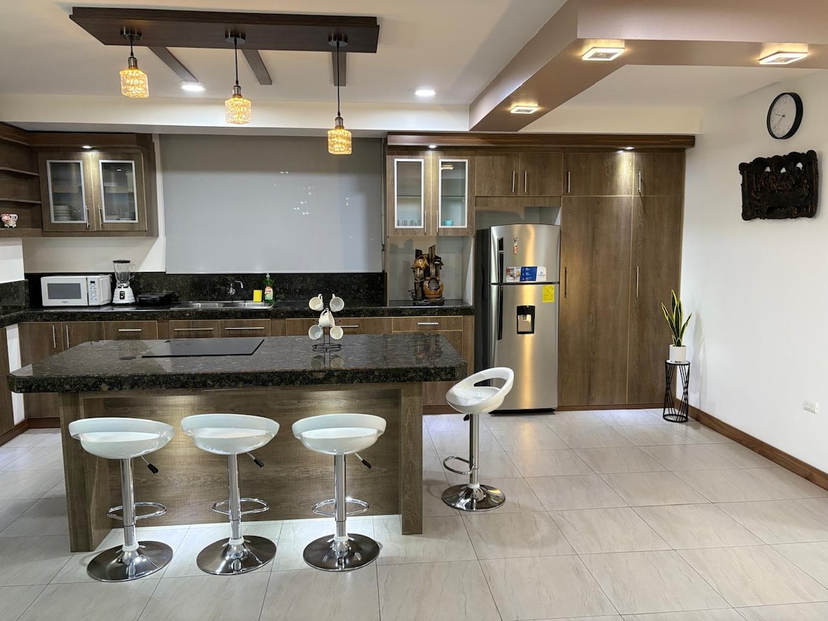 A modern kitchen area features sleek wooden cabinets and a spacious countertop. Three white bar stools are positioned at the kitchen island, which is illuminated by pendant lights. A stainless steel refrigerator stands against the wall, complemented by appliances on the countertop.