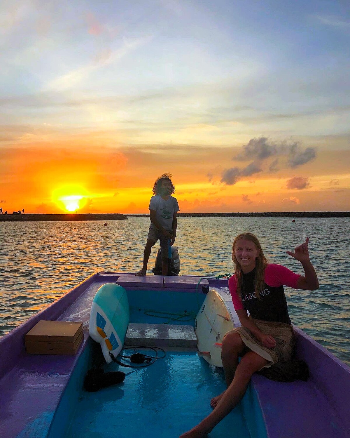 An image capturing two individuals on a boat during sunset, with vibrant colors reflecting on the water. One person stands at the bow, while the other sits comfortably, giving a casual hand gesture. The horizon is adorned with soft clouds against a warm, glowing sky.