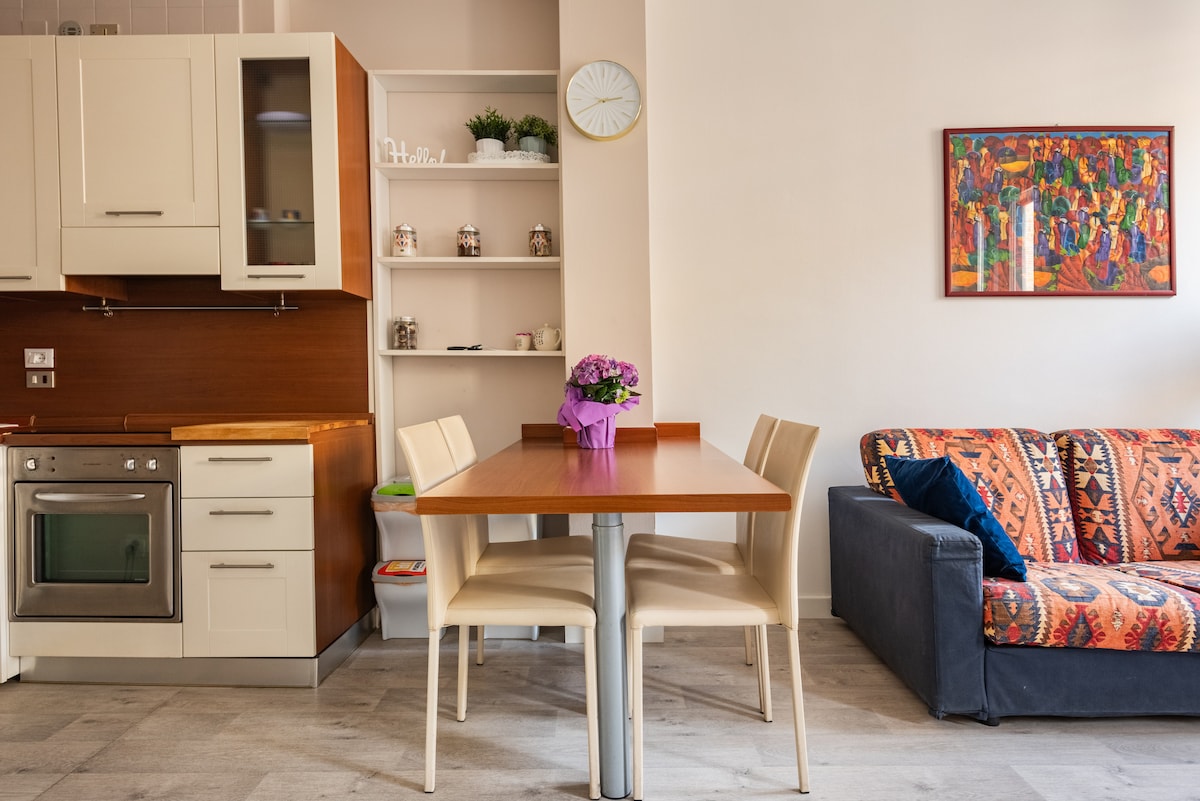 A modern kitchen is visible with sleek cabinetry and an oven. A wooden dining table is set for four, complemented by light-colored chairs. A vibrant floral arrangement centers the table. A cozy sofa is dressed in colorful, patterned fabric, adding warmth to the living space.